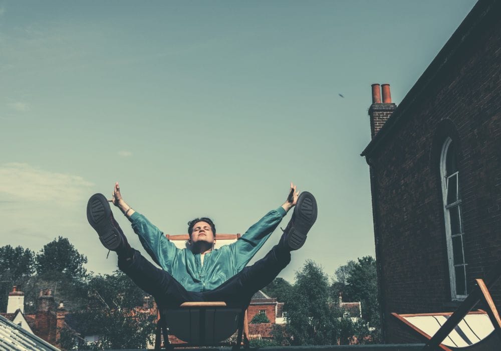 Luke, sitting in a deckchair on a rooftop terrace, with his eyes closed and his arms and legs raised.