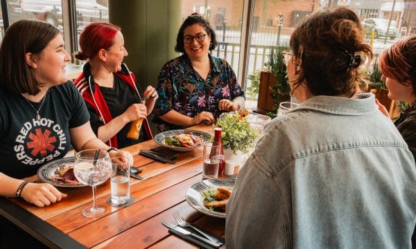 A number of people sit around a wooden table with food and drinks socialising at No 60 ARC's café bar.