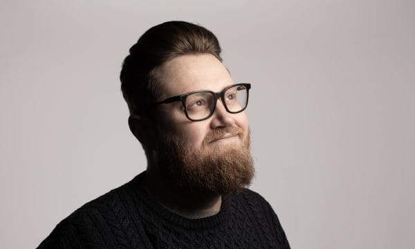 A headshot of comedian Pete Selwood, a white man with short brown hair, black rimmed glasses, and a brown beard. He is looking off to the right, and is smiling.