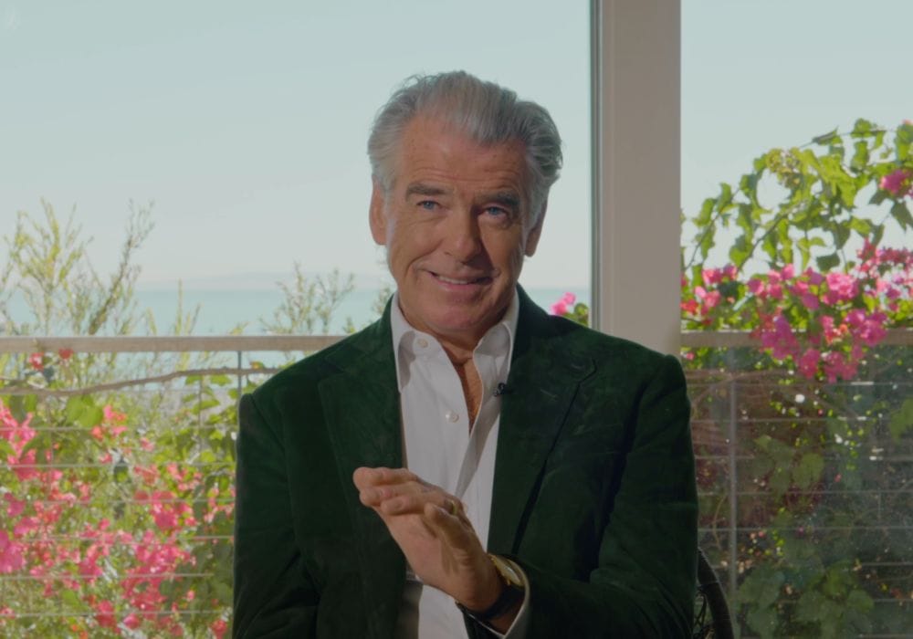 Headshot of Pierce Brosnan a white male with grey hair wearing a blue suit with white shirt.