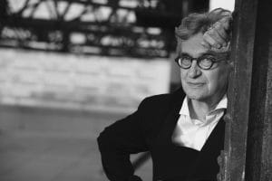 Black & White image of Wim Wenders a white male with grey hair wearing black glasses. He has a white shirt and Black jacket.