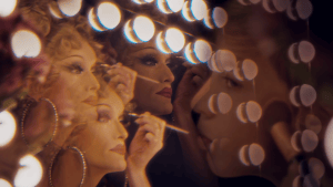 A drag queen looking in the mirror to apply makeup with a young boy looking back. 