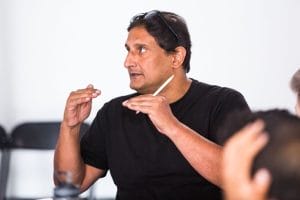 Ishy Din, wearing a black round-necked t-shirt and glasses pushed back on top of his head, is holding a pen in one hand and is speaking to a group of people (out of shot)