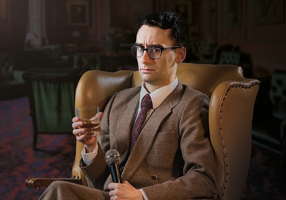 MC Hammersmith, a white man wearing glasses and a suit and tie, sits in a large, leather armchair. In one hand is a tumbler of whisky, in the other he holds a microphone.