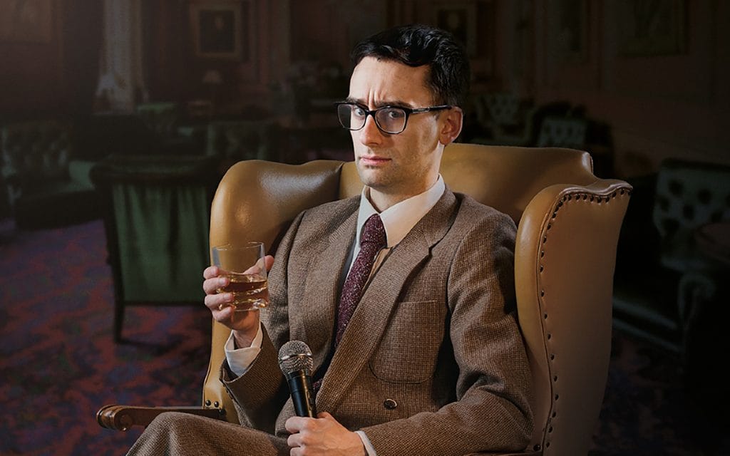 MC Hammersmith, a white man wearing glasses and a suit and tie, sits in a large, leather armchair. In one hand is a tumbler of whisky, in the other he holds a microphone.