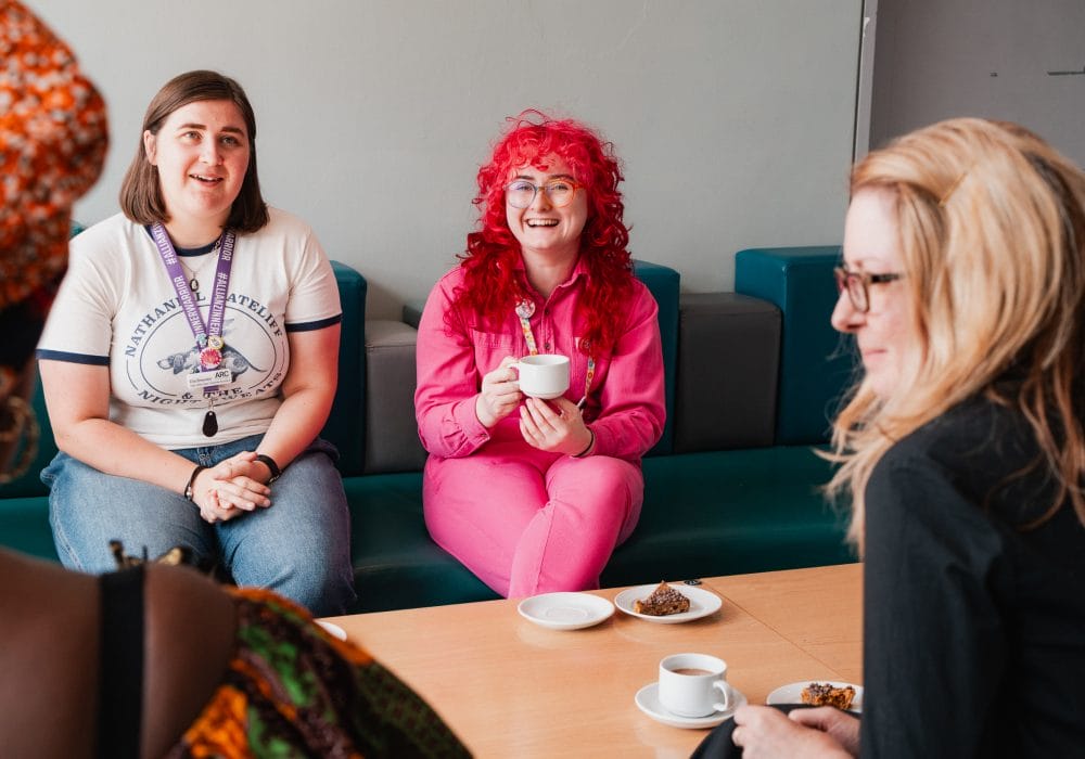 Several people sitting around a table, chatting, drinking coffee, and eating cake. The photo focuses on Allison, one of ARC's Producers. She is wearing a bright pink jumpsuit and has bright pink hair.