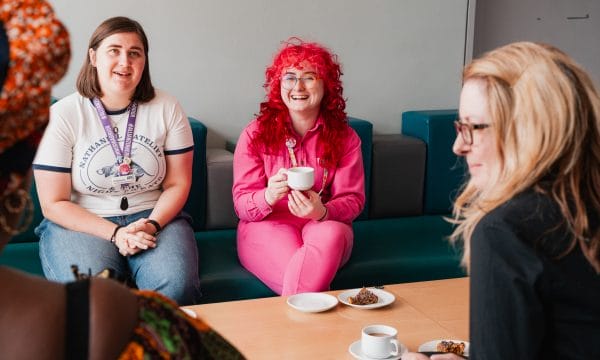 Several people sitting around a table, chatting, drinking coffee, and eating cake. The photo focuses on Allison, one of ARC's Producers. She is wearing a bright pink jumpsuit and has bright pink hair.