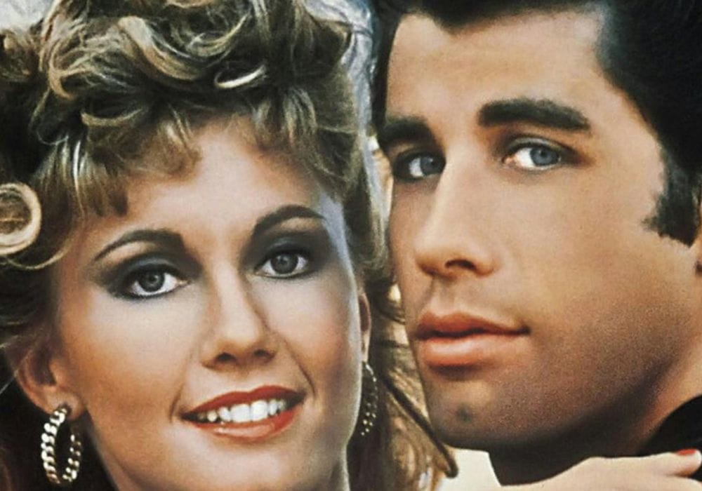 A close-up of Olivia Newton-John and John Travolta. Olivia Newton-John is looking directly at the viewer, with John Travolta looking at the viewer over his shoulder.