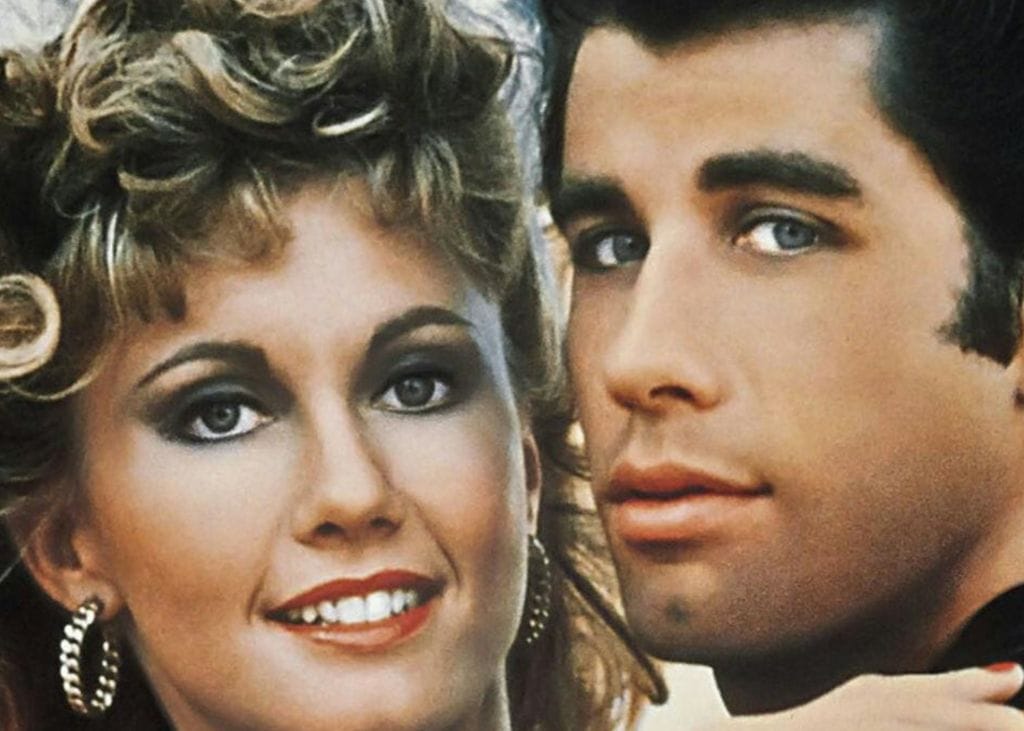 A close-up of Olivia Newton-John and John Travolta. Olivia Newton-John is looking directly at the viewer, with John Travolta looking at the viewer over his shoulder.