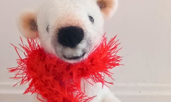 A close-up of a needle felt polar bear, he has light pink ears and nose, and is wearing a bright red fuzzy scarf.