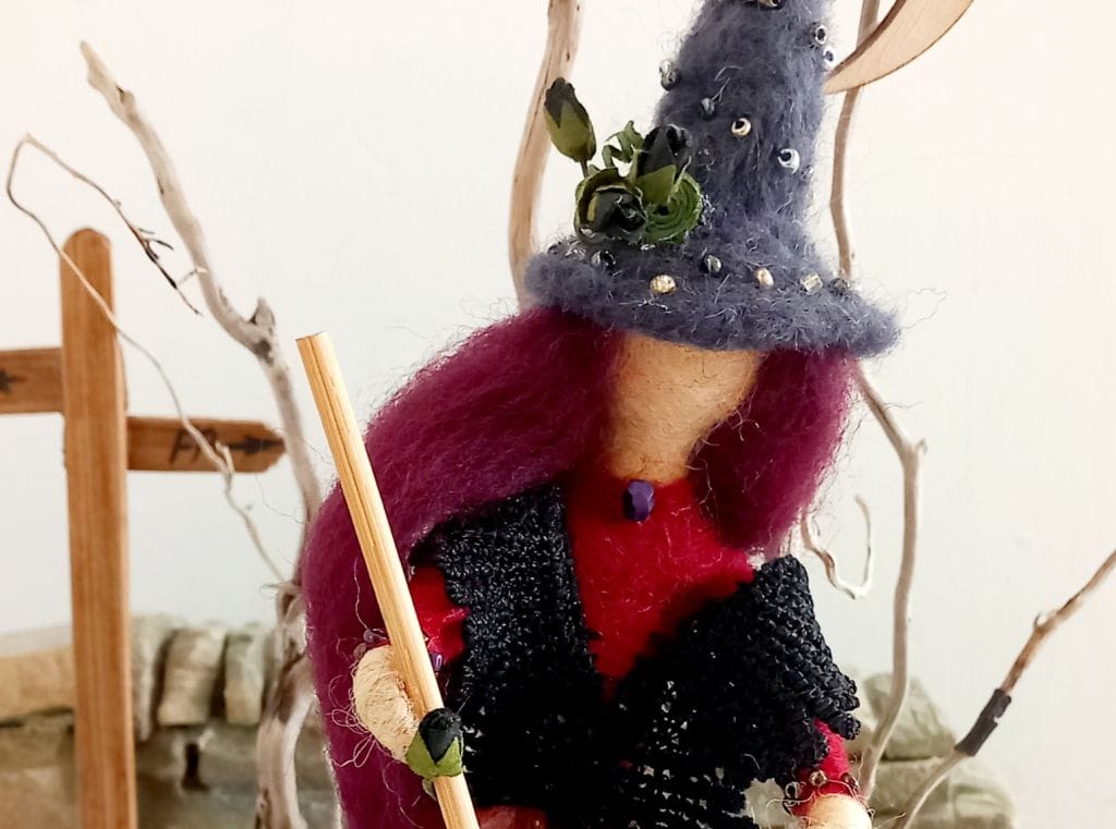 A needle felt sculpture of a witch, she has long purple hair, a black beaded shawl, and a tall grey hat decorated with leaves.