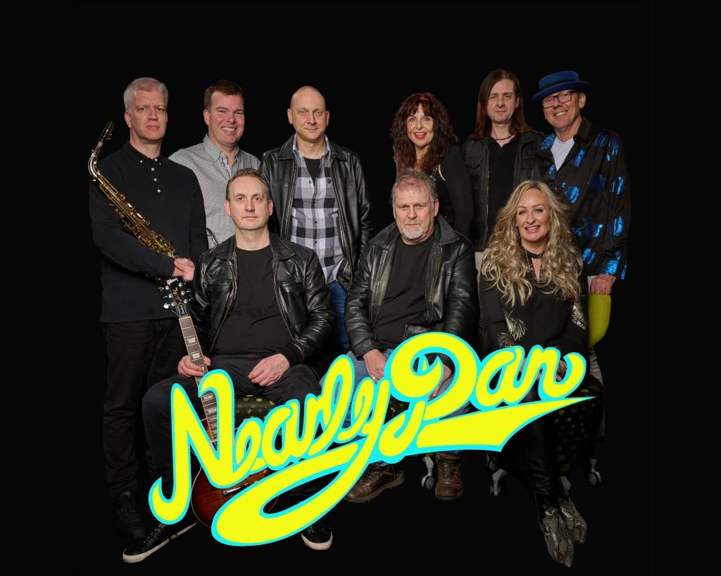 The image shows a group portrait of a band called "Nearly Dan." The band members include a saxophonist, a guitarist, a keyboardist, a female vocalist, and several other musicians. They are posed in front of a large "Nearly Dan" logo in vibrant yellow and blue colors. The members are dressed in a mix of casual and more formal attire, suggesting a professional music group. The image has a dark background, drawing focus to the band members and the logo.