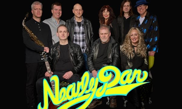 The image shows a group portrait of a band called "Nearly Dan." The band members include a saxophonist, a guitarist, a keyboardist, a female vocalist, and several other musicians. They are posed in front of a large "Nearly Dan" logo in vibrant yellow and blue colors. The members are dressed in a mix of casual and more formal attire, suggesting a professional music group. The image has a dark background, drawing focus to the band members and the logo.