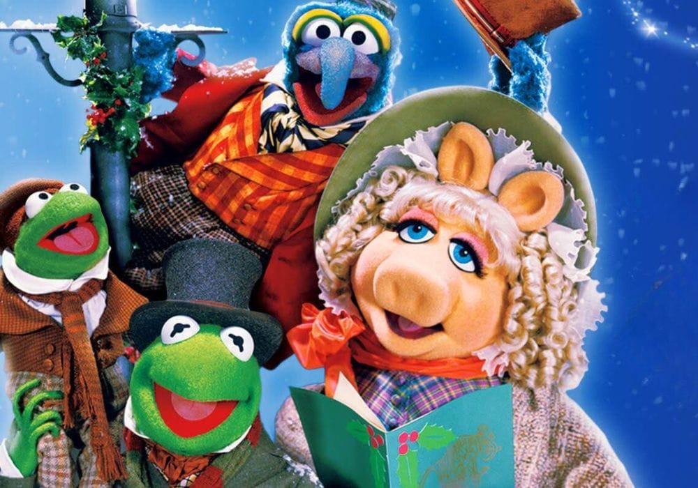 The Muppets puppets Kermit the Frog, Miss Piggy, and Gonzo are all crowded around a lamppost dressed in Victorian style clothing. Miss Piggy is holding a songbook decorated with holly leaves, Gonzo is tipping his hat, and Kermit the Frog is holding a smaller frog on his shoulder.