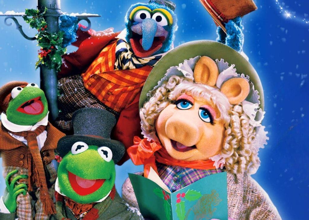 The Muppets puppets Kermit the Frog, Miss Piggy, and Gonzo are all crowded around a lamppost dressed in Victorian style clothing. Miss Piggy is holding a songbook decorated with holly leaves, Gonzo is tipping his hat, and Kermit the Frog is holding a smaller frog on his shoulder.