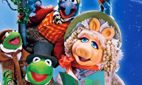 The Muppets puppets Kermit the Frog, Miss Piggy, and Gonzo are all crowded around a lamppost dressed in Victorian style clothing. Miss Piggy is holding a songbook decorated with holly leaves, Gonzo is tipping his hat, and Kermit the Frog is holding a smaller frog on his shoulder.