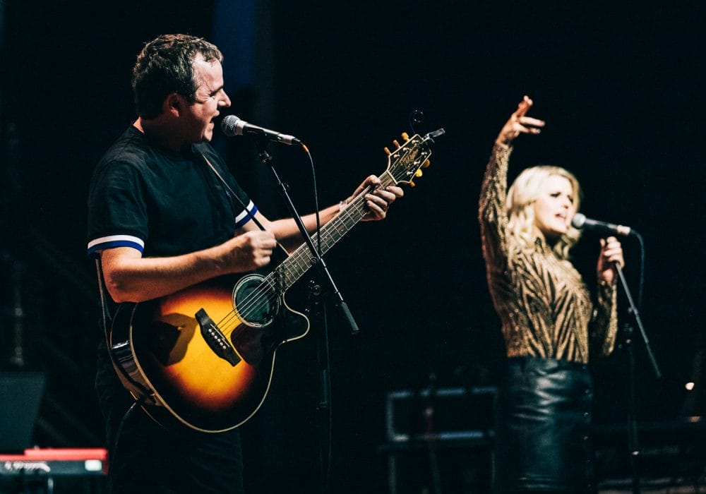 A live music performance on stage. In the foreground, there's a man playing an acoustic guitar and singing into a microphone. He's wearing a dark shirt with light-colored stripes on the sleeves. To his right, there's a woman with long blonde hair singing into another microphone, with her arm raised expressively. In the background on the left, there's a person seated at a keyboard, which appears to be a Nord brand synthesizer based on its red color. The lighting creates a dramatic atmosphere, with the performers illuminated against a dark background. The guitarist's instrument appears to be a sunburst-colored acoustic-electric guitar. The overall scene captures the energy and collaboration of a live band performance, with multiple musicians contributing to the sound.