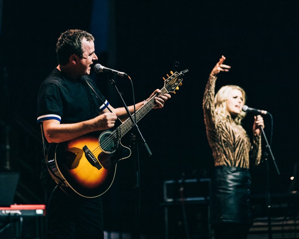 A live music performance on stage. In the foreground, there's a man playing an acoustic guitar and singing into a microphone. He's wearing a dark shirt with light-colored stripes on the sleeves. To his right, there's a woman with long blonde hair singing into another microphone, with her arm raised expressively. In the background on the left, there's a person seated at a keyboard, which appears to be a Nord brand synthesizer based on its red color. The lighting creates a dramatic atmosphere, with the performers illuminated against a dark background. The guitarist's instrument appears to be a sunburst-colored acoustic-electric guitar. The overall scene captures the energy and collaboration of a live band performance, with multiple musicians contributing to the sound.