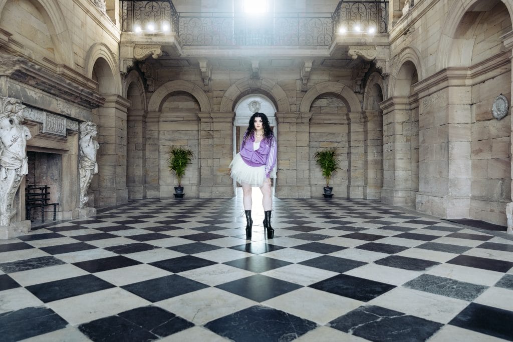 Image of a woman in a tracksuit, tutu, and black boots stood inside a grand stone building.