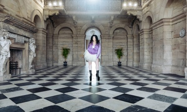 Image of a woman in a tracksuit, tutu, and black boots stood inside a grand stone building.