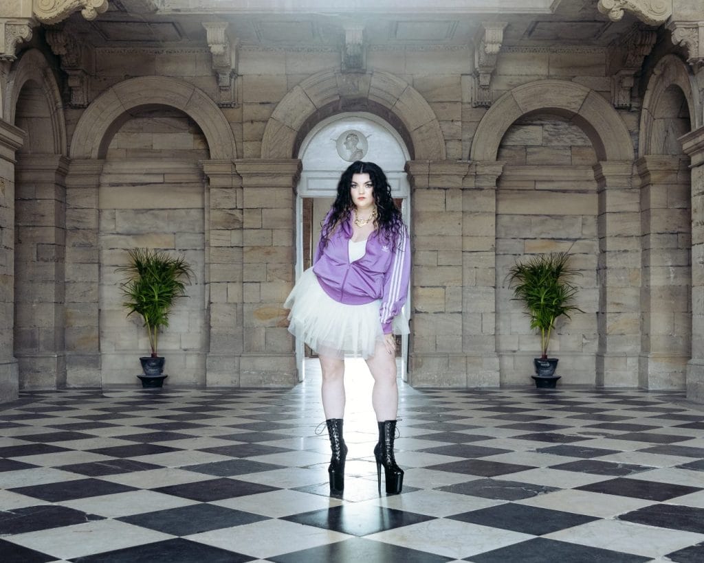 Image of a woman in a tracksuit, tutu, and black boots stood inside a grand stone building.