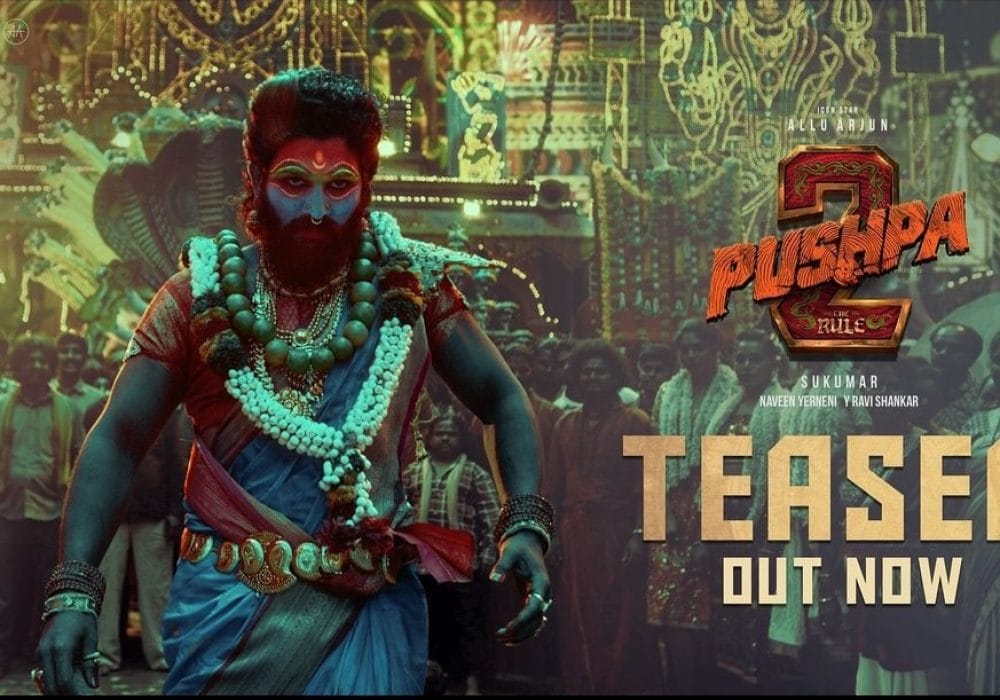 The actor Allu Arjun dressed in ornate clothing, with his face painted blue, red, and gold. Behind him is a crowd gathered in front of a building with intricate painted patterns on it. To the right of the image are the words Pushpa 2: The Rule