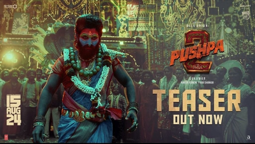 The actor Allu Arjun dressed in ornate clothing, with his face painted blue, red, and gold. Behind him is a crowd gathered in front of a building with intricate painted patterns on it. To the right of the image are the words Pushpa 2: The Rule