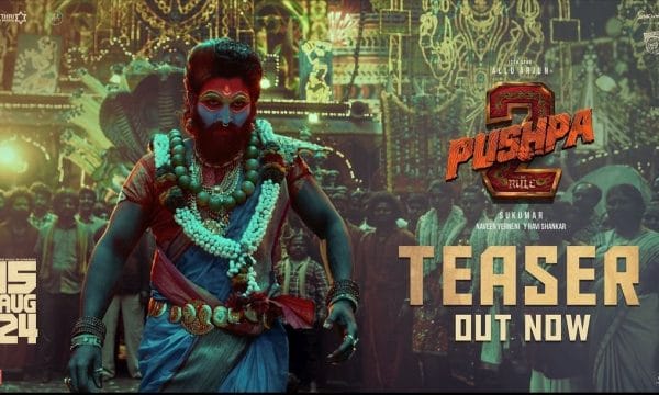 The actor Allu Arjun dressed in ornate clothing, with his face painted blue, red, and gold. Behind him is a crowd gathered in front of a building with intricate painted patterns on it. To the right of the image are the words Pushpa 2: The Rule