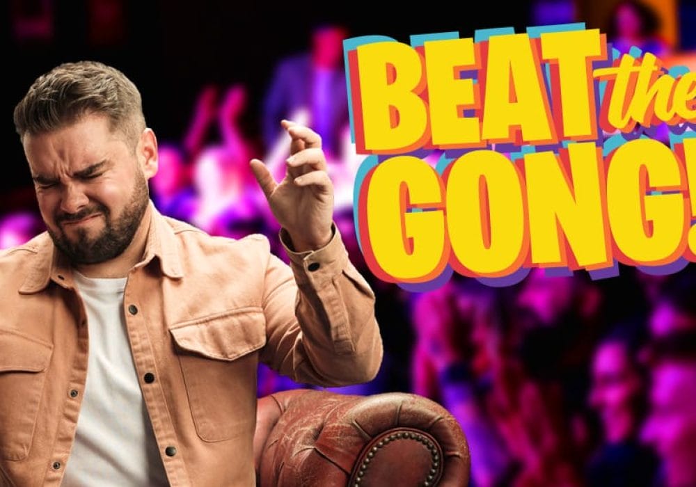Comedian Adam Rowe, a white man with short dark hair, and a short dark beard, is grinning broadly while looking at the camera. To his right is the Beat the Gong logo.