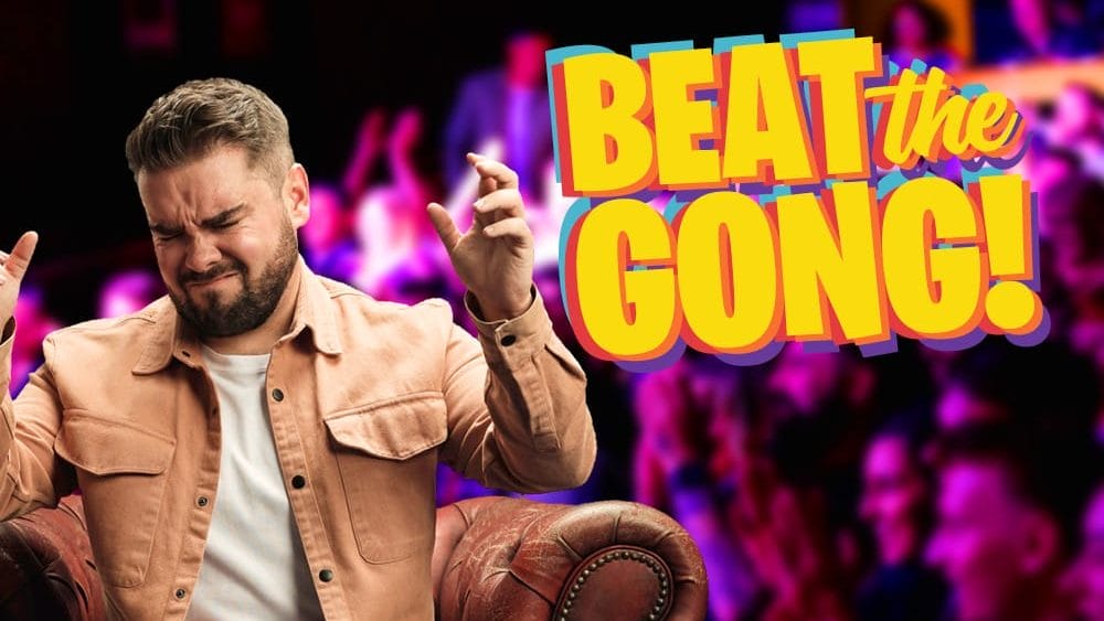Comedian Adam Rowe, a white man with short dark hair, and a short dark beard, is grinning broadly while looking at the camera. To his right is the Beat the Gong logo.