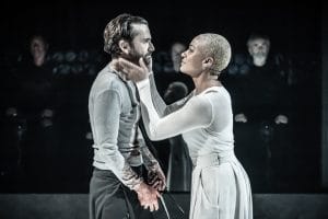 Side profiles of David Tennant a white male with dark hair who is wearing a grey long-sleeved t-shirt holding a knife in his left hand. Cush Jumbo a mixed race female with short blonde hair is wearing a white dress and has David Tennant's face in her hands.