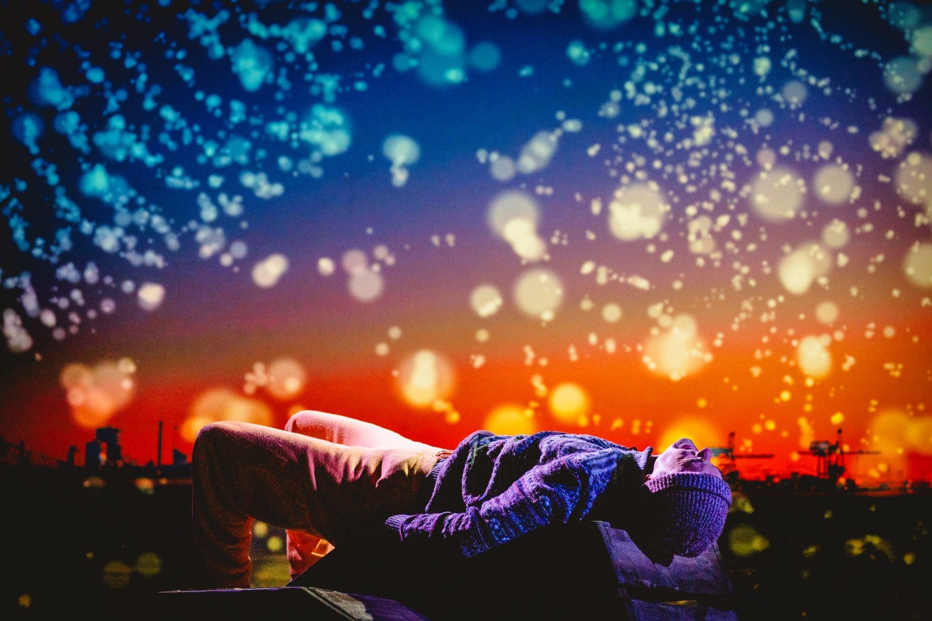 An actor wearing a knitted beanie and thick knitted jumper lays staring at the sky. The background is orange and blue, filled with flecks of light. Factories are silhouetted along the horizon.