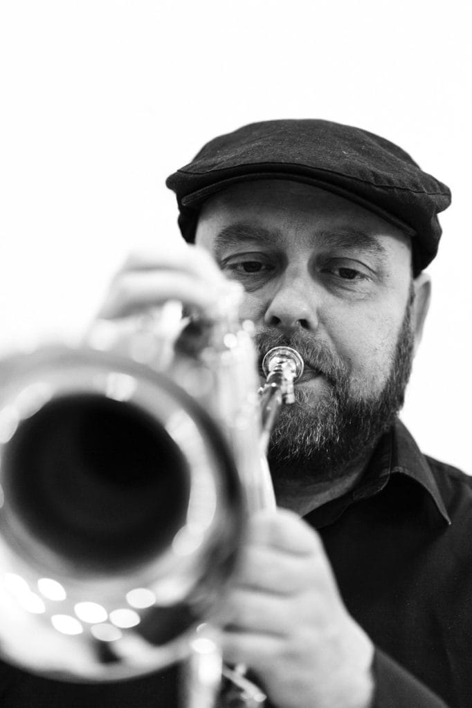 a black and white photograph of a bearded man wearing a flat cap and dark shirt, playing what appears to be a trumpet or cornet. The image has an artistic quality with nice depth of field - the musician is in sharp focus while the background is softly blurred. The composition draws attention to both his concentrated expression and the brass instrument he's playing.