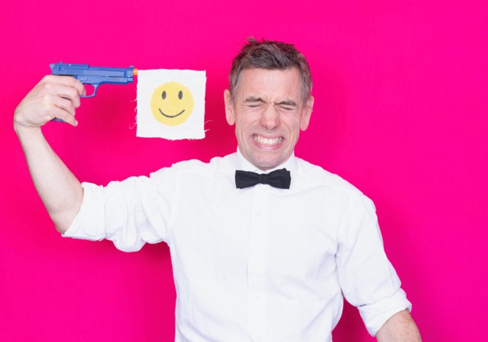 Actor Dan Bye stands against a neon pink background, wearing a white shirt and a black bowtie. He is holding a blue toy gun to his head, and a smiley face flag hangs from the end of it. He is grimacing.