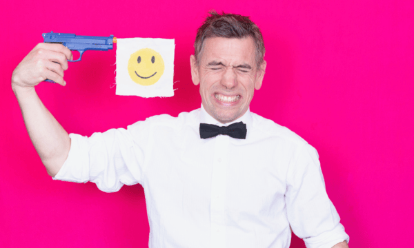 Actor Dan Bye stands against a neon pink background, wearing a white shirt and a black bowtie. He is holding a blue toy gun to his head, and a smiley face flag hangs from the end of it. He is grimacing.