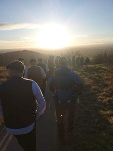 Photograph of members of Infant Hercules men's choir walking down a country road with the sunning setting in the far distance creating a lens flare effect across the image.