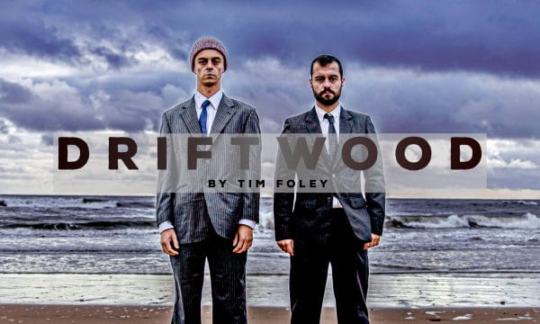 Two men wearing dark suits stand on a beach, above them are grey clouds. Text reads Driftwood.