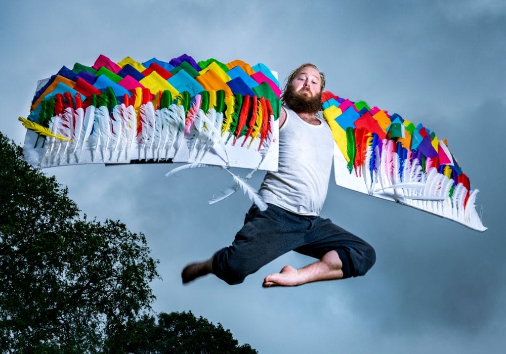 Actor James Rowland leaps in front of a cloudy, grey sky. He has his arms outstretched wearing wings decorated with colourful fabric.