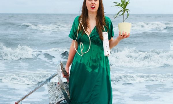 An actress stands in front a choppy sea, she is wearing a long green dress. In one hand she holds a potted plant, in the other a metal stepladder. She has a white extension cord draped over her shoulders.