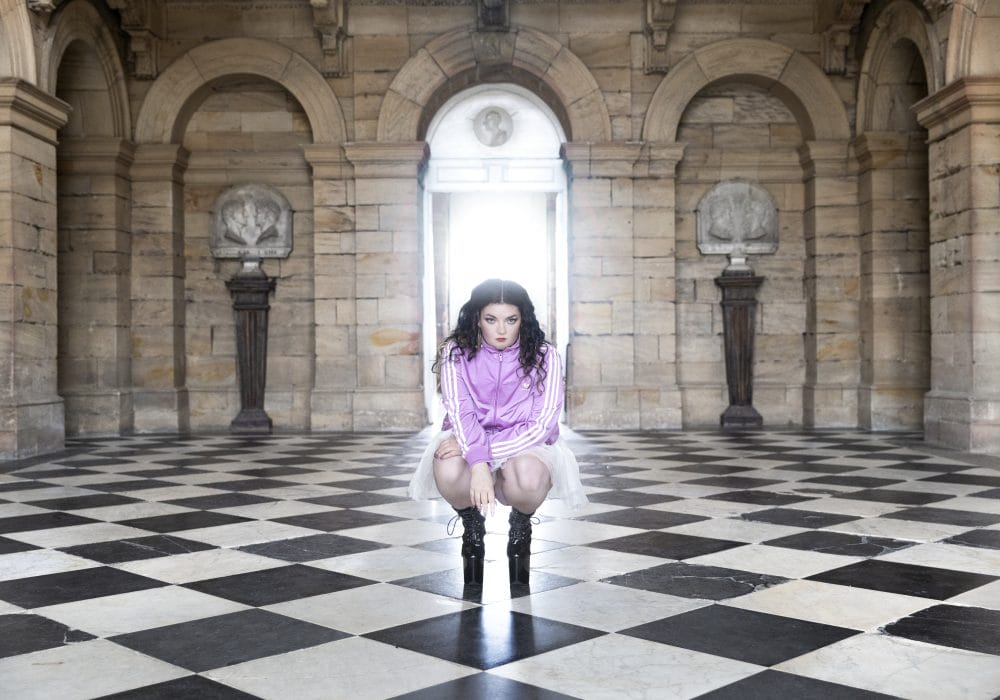 Image of a woman in a tracksuit, tutu, and black boots stooped inside a grand stone building.