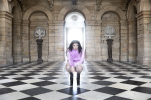 Image of a woman in a tracksuit, tutu, and black boots stooped inside a grand stone building.