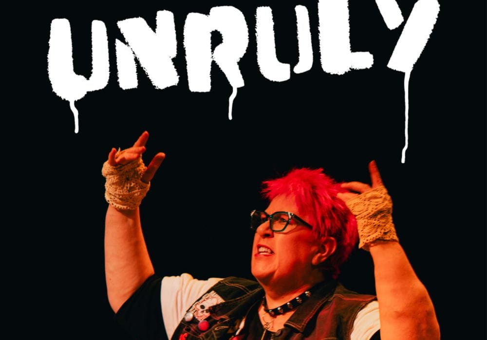 Actress Vici Wreford-Sinnot, with short, bright pink hair, raises her arms up, she has a defiant expression. Above her is the word Unruly, in white spray paint.