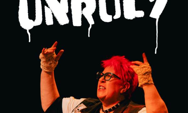 Actress Vici Wreford-Sinnot, with short, bright pink hair, raises her arms up, she has a defiant expression. Above her is the word Unruly, in white spray paint.