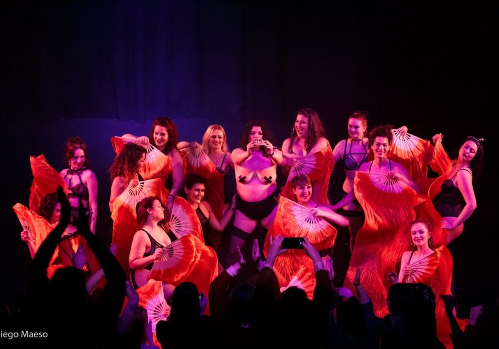 A group of burlesque performers stand in the centre of a stage holding various poses.