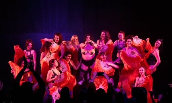 A group of burlesque performers stand in the centre of a stage holding various poses.