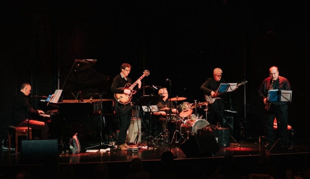 A five piece jazz band perform on stage to a crowd