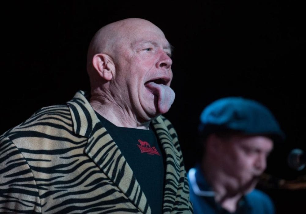 Buster Bloodvessel on stage in his typical pose with his tongue out. Another musician alongside him is blurred in the background.