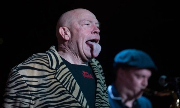 Buster Bloodvessel on stage in his typical pose with his tongue out. Another musician alongside him is blurred in the background.