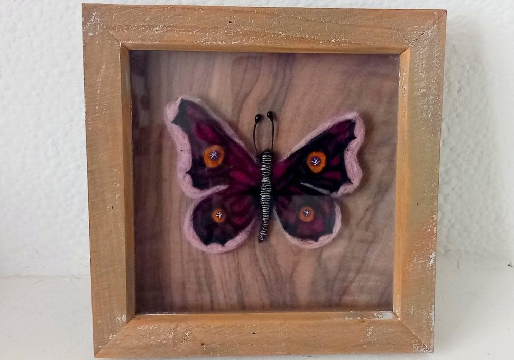 A pink and yellow butterfly made from needle felting. It has an intricate pattern on it's wings, and it's body is black with white stripes. It is in the centre of a wooden frame.