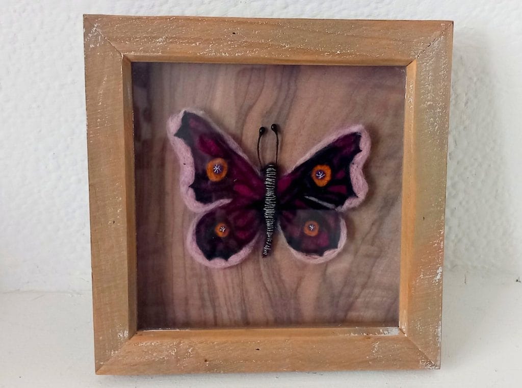 A pink and yellow butterfly made from needle felting. It has an intricate pattern on it's wings, and it's body is black with white stripes. It is in the centre of a wooden frame.